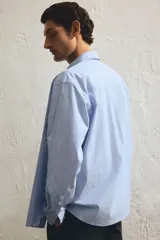 Camisa celeste y blanca a rayas verticales, de corte holgado, manga larga, cuello inglés, bolsillo en el pecho y cierre con botones.