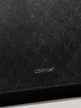 Cartera de mano Calvin Klein con correa ajustable para el hombro. El material exterior es de piel sintética negra con un patrón texturizado de monograma repetido. Presenta una solapa con cierre magnético y un logo metálico plateado en la parte frontal.