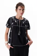Blusa negra de algodón con bordados blancos a contratono, cuello redondo con lazo y borlas decorativas, y mangas cortas.