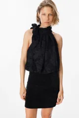 Blusa blanca sin mangas con cuello halter y lazo lateral. Confeccionada en tela de gasa con textura floreada en relieve.