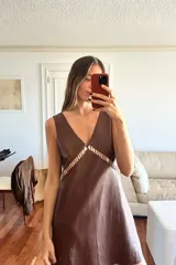 Vestido corto de cuero ovino color marrón, con escote en V profundo y detalle de pasamanería decorativa en el frente. Presenta un corte holgado y diseño minimalista sin mangas.