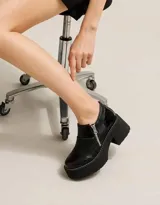 Zapatos de tacón alto y plataforma con diseño de botineta baja en color negro, confeccionados con una combinación de cueros con diferentes texturas, costuras pespunteadas en blanco y cierre metálico lateral.