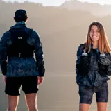 Mochila chaleco de hidratación Merrell en color negro, diseñada para running y trail running, con múltiples bolsillos frontales, cordones de ajuste y tejido de malla transpirable.