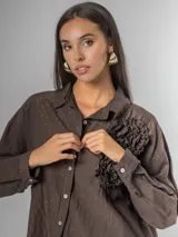 Camisa de manga larga con cuello clásico, confeccionada en tela nacarada con brillo sutil. Presenta apliques de flores tridimensionales fruncidas en el frente, una en color beige claro y dos en color marrón oscuro.