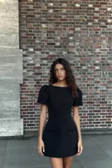Vestido corto negro con cuello barco, mangas cortas abullonadas y pliegues. Presenta falsos bolsillos de vivo en el delantero y costuras marcadas. Cierre con cremallera oculta en la espalda.