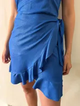 Conjunto de dos piezas en lino con lurex color azul brillante. Incluye una falda corta cruzada con volados y un top halter con escote recto y lazo ajustable en el contorno.