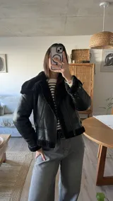 Campera tipo gamulán de cuero sintético negro con interior y detalles de cuello y puños en piel sintética negra. Presenta cierre frontal, correas ajustables en los puños y diseño de corte clásico.