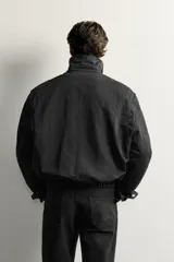 Cazadora negra de corte regular fit, confeccionada en tejido técnico con mezcla de algodón. Cuenta con cuello solapa, manga larga con puño abotonado, bolsillos de vivo en la cadera, pliegues laterales en la espalda y bajo elástico. Cierre frontal con cremallera.