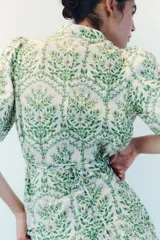Vestido largo de algodón color blanco con estampado botánico en tonos verdes. Presenta cuello mao, cierre frontal con botones ocultos, mangas cortas abullonadas y falda de corte escalonado con volados. Incluye un cinturón fino para ajustar en la cintura.