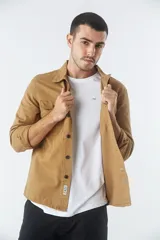 Sobrecamisa de jean color tostado, con cuello clásico, cierre frontal con botones y dos bolsillos frontales con solapa. Presenta mangas largas con puños abotonados y un pequeño parche decorativo en el ruedo.