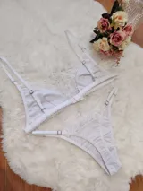 Conjunto de lencería compuesto por un bralette triangular y una tanga, ambos confeccionados en algodón blanco con detalles de encaje en los bordes. El bralette cuenta con breteles ajustables y la tanga presenta laterales regulables.
