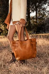 Cartera de cuero color marrón tostado, con doble asa de hombro y diseño de hebillas decorativas en los costados. Incluye doble bolsillo exterior y colgante interno para llaves.