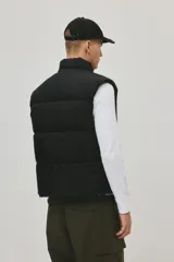 Chaleco acolchado negro de nylon guateado, repelente al agua y rompevientos. Cuenta con cuello elevado, cierre bidireccional con protección para el mentón, bolsillos laterales con cierre y un bolsillo interior con botón a presión. El bajo tiene un cordón elástico con topes para ajustar.