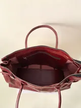 Bolso grande de cuero marrón con textura tipo cocodrilo, estructura rígida y asas para llevar al hombro. Tiene una tira con tachas y hebillas en cada extremo, también lleva lazos en los costados.