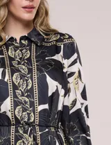 Vestido corto de satén estampado en tonos negro y beige con diseño floral y geométrico, cuello camisero con solapa, mangas largas con puños, lazo ajustable en la cintura y falda con volado en el ruedo.