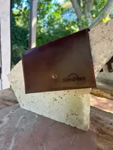 Sobre de cuero color marrón, con cierre de botón a presión en solapa. Presenta costuras visibles en los bordes y el logo grabado en la solapa.