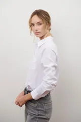 Camisa blanca de manga larga con cuello clásico y botones frontales. Tiene un bolsillo en el pecho con un pequeño logo bordado.