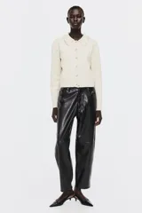 Saco corto de punto suave color beige, con cuello camisero con ribete de volados y botones de metal con diseño de nudo. Tiene hombros ligeramente caídos y remate acanalado en puños y bajo.