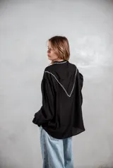 Camisa negra de manga larga con bordados blancos en la parte superior delantera y hombros. Presenta cuello mao, cierre frontal con botones y mangas con puños ajustados.
