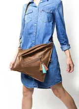 Cartera bandolera de cuero color marrón claro, con correa larga regulable de cuero marrón oscuro. Tiene cierre con solapa y broche imantado, bolsillo frontal con cierre y detalle de borlas decorativas de colores.