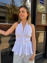 Blusa halter blanca sin mangas, con escote en V pronunciado, espalda descubierta, cintura fruncida con doble lazo frontal ajustable y diseño peplum con vuelo en la parte inferior.