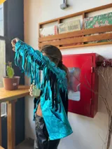 Campera turquesa de tela brillante con flecos en mangas y ruedo.