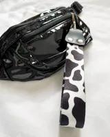 Llavero de muñeca con cinta de tela estampada con diseño de vaca en blanco y negro. Tiene un conector de cuero sintético negro con remaches plateados y un gancho metálico plateado.