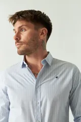 Camisa de hombre de corte slim fit, confeccionada en algodón con diseño de rayas verticales finas en tonos claros. Presenta cuello clásico, cierre frontal con botones y logo bordado en el pecho.