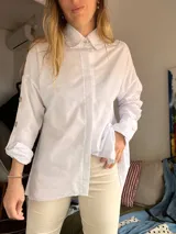 Camisa blanca de popelina de algodón, de corte oversize, con cuello camisero, manga larga y detalles de botones decorativos en las mangas.