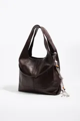 Bolso de mano y hombro grande de cuero marrón oscuro, tipo hobo, con diseño estructurado mediante costuras frontales y asa integrada. Incluye detalle lateral de cordones blancos con cuentas decorativas y el logotipo plateado de la marca en la parte inferior frontal.