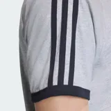Remera de algodón gris con cuello redondo y bordes de mangas en color negro. Presenta las icónicas tres tiras de la marca en los hombros y el logo del trébol bordado en el pecho.