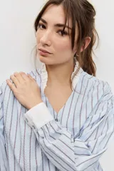 Camisa de rayas celestes y marrones, de corte holgado y manga larga, con cuello en V y detalles blancos en el cuello y los puños.
