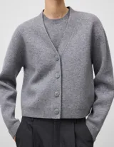 Cárdigan corto de lana cocida en color gris melange, con cuello en V, manga larga y cierre frontal mediante botones. Presenta terminaciones en punto canalé en puños y bajo.