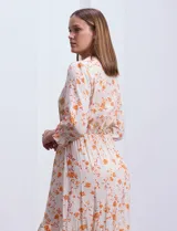 Vestido midi de gasa con estampado geométrico y floral en tonos naranja y turquesa sobre base beige. Presenta escote en V, cierre frontal con botones, mangas 3/4 con puños fruncidos y cintura elastizada con lazo de ajuste. El diseño incluye un ruedo con volado y corte asimétrico más largo en la parte posterior.