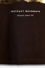 Camiseta de algodón color marrón oscuro, de corte regular, cuello redondo y manga corta. Presenta una estampación metalizada dorada en el parte frontal con texto "REYKJAVIK ART GALLERY" y "ABSTRACT MOVEMENTS Reykjavik, Iceland, 1975".