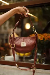 Cartera pequeña de cuero marrón con diseño semicircular, detalles metálicos de bronce y asa ajustable con ganchos.