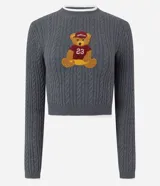 Sweater cropped de punto gris con diseño trenzado, cuello redondo, puños y ruedo con ribetes blancos. Presenta un aplique bordado en el pecho con diseño de osito deportista.
