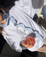 Camisa de manga larga abullonada en color rosa con rayas verticales finas. Presenta un diseño con volados en el cuello y a lo largo de la botonadura frontal, puños con detalle de rayas contrastantes y botones decorativos dorados.