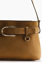 Cartera de hombro color tostado con acabado de gamuza, correa ajustable y tira decorativa frontal con hebilla metálica dorada.