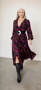 Vestido midi de seda natural con estampado floral en tonos fucsia sobre base negra. Presenta un diseño drapeado con escote en V cruzado, mangas largas abullonadas y una abertura frontal en la falda.