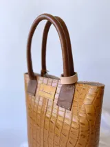 Bolso matero de cuero color beige con textura de reptil y detalles en cuero marrón con textura de cocodrilo. Incluye manijas cortas de cuero marrón y dije de cruz plateada.