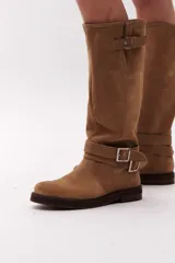 Bota de caña alta color marrón chocolate, confeccionada en gamuza. Presenta dos correas desmontables con hebillas metálicas plateadas.