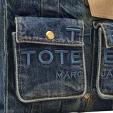 Bolso tipo tote de Marc Jacobs confeccionado en denim, con diseño estructurado que incluye dos bolsillos frontales con solapa y cierre de broche metálico. Presenta el logo bordado en el frente y asas superiores reforzadas junto con una correa ajustable para llevar al hombro.