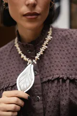 Collar corto con dije central en forma de hoja, elaborado en guampa con engarce de alpaca. La cadena está compuesta por bastoncitos de acrílico y mostacillas transparentes, finalizando con una cadena de eslabones de acero quirúrgico ajustable.