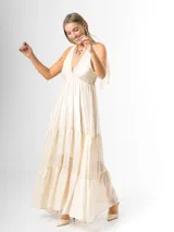 Vestido largo color crudo, confeccionado en mezcla de tencel y lino, con escote en V pronunciado, breteles anchos terminados en lazos en los hombros, y falda larga de varios niveles con volados amplios y fluidos.