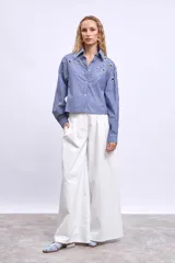 Camisa de mangas largas a rayas verticales azules y blancas, con cuello clásico y cierre frontal con botones. Presenta apliques de strass y ojalillos metálicos.