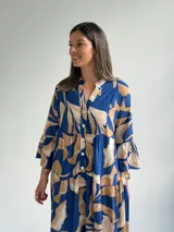 Vestido largo azul con estampado de hojas en tonos beige y blanco, escote en V con botones frontales, mangas largas amplias y diseño escalonado.