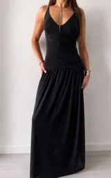 Vestido largo negro con escote profundo en V, tirantes finos y diseño ajustado en la cintura con detalle fruncido, que se abre en una falda fluida y amplia.