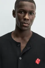 Cárdigan de punto negro, corte relajado, con cuello redondo, manga larga y cierre frontal de botones oscuros. Presenta un parche bordado en forma de dado rojo con corazones blancos en el lado izquierdo del pecho.