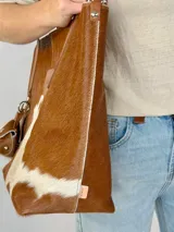 Bolso tipo tote Chabas modelo Molly, confeccionado en cuero con pelo natural en tonos marrón y blanco. Posee asas largas de cuero marrón, parche frontal con la marca grabada y un charm colgante decorativo en forma de estrella con detalle de pelo.
